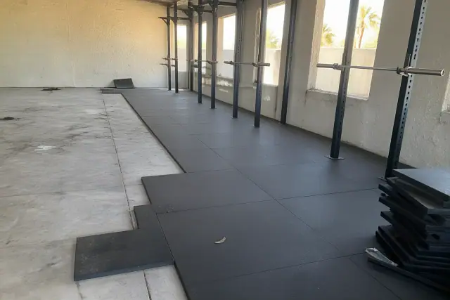 Technician installing heavy-duty rubber floor tiles in a Phoenix, AZ gym with squat racks and concrete walls.