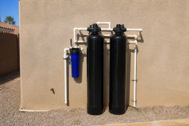 Carbon filtration system tanks with PVC piping on a tan stucco wall at a house in Tempe, AZ.